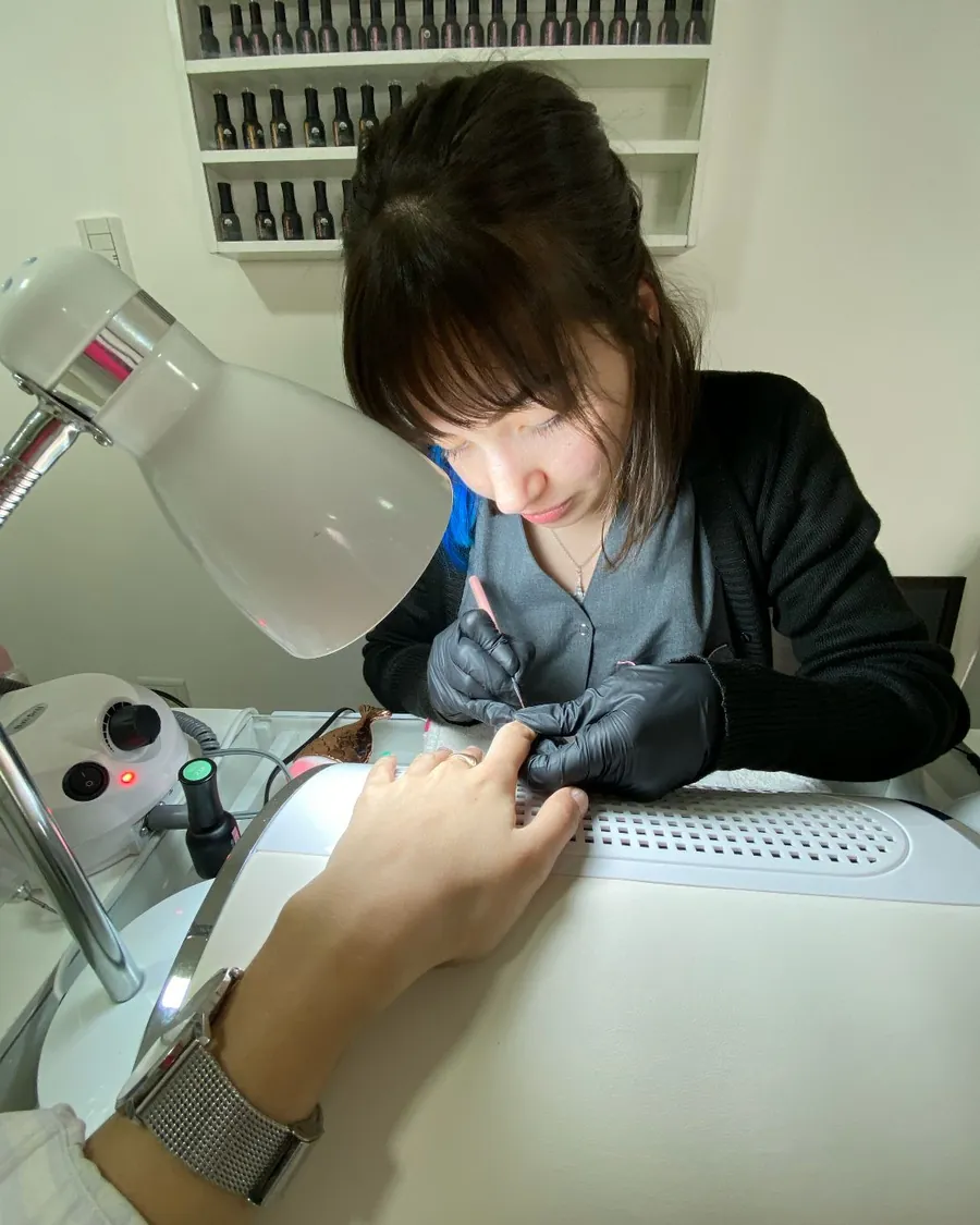 Erica at a salon table in black gloves, painting a client's nails under the lamp